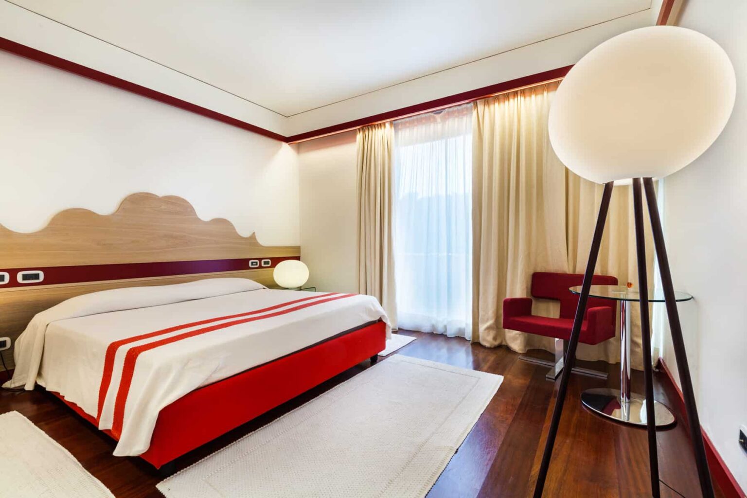 Modern hotel bedroom featuring a red and white color scheme, a comfortable bed with decorative linens, a stylish floor lamp, a cozy chair, and large windows with sheer curtains allowing natural light to fill the space. | Only on SopranoVillas