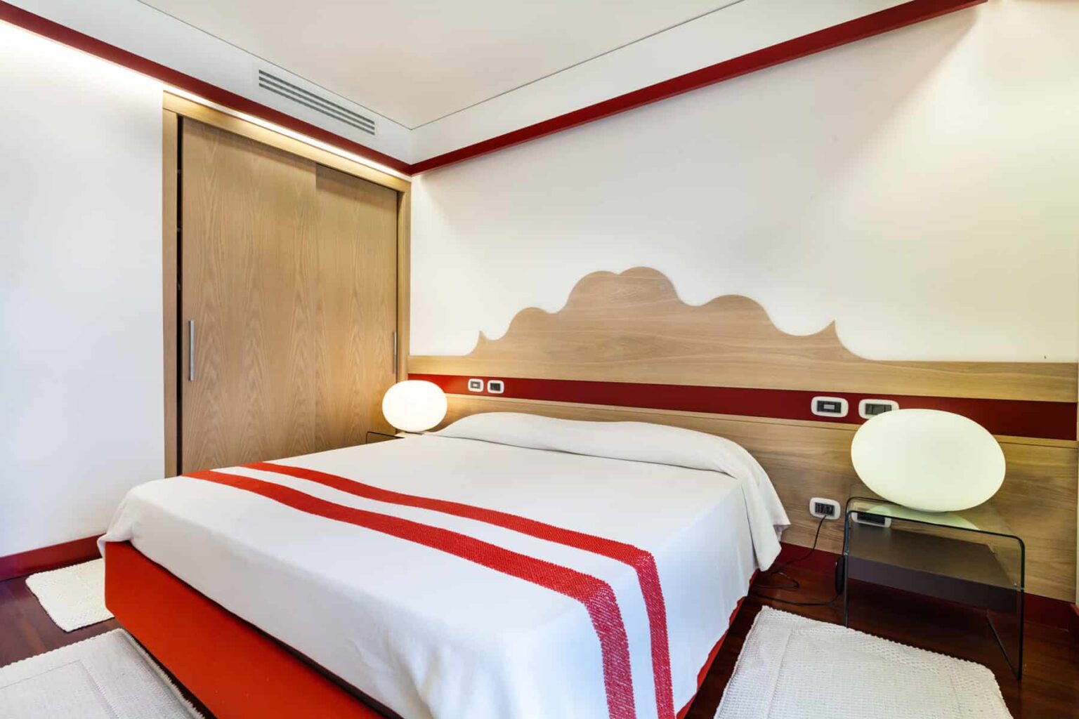 Modern hotel room featuring a queen-sized bed with a stylish red and white color scheme, minimalist decor, and contemporary lighting. The room includes a wooden wardrobe and a sleek side table, creating a cozy and inviting atmosphere. Ideal for travelers seeking comfort and style. | Only on SopranoVillas