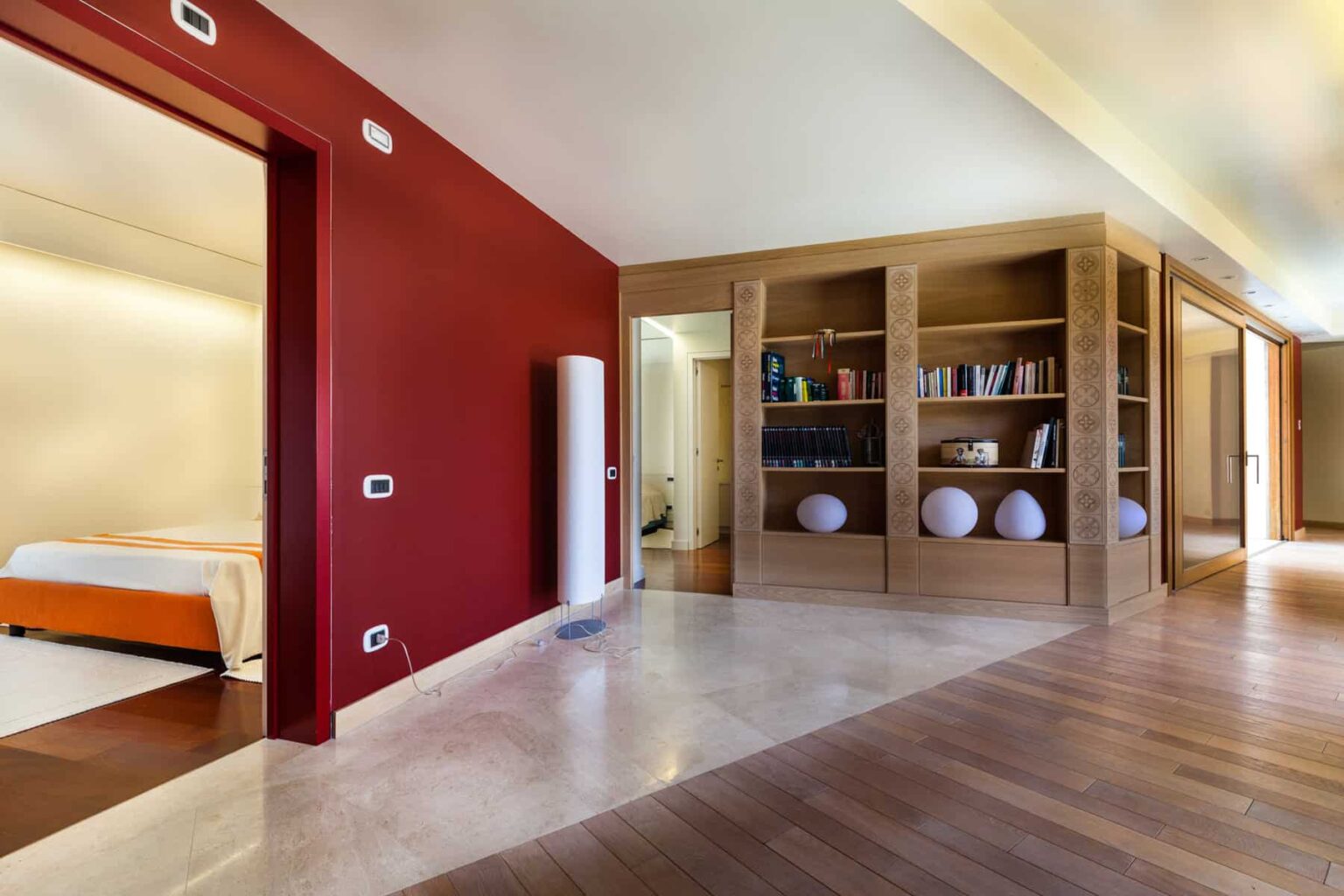 Modern interior design showcasing an open living space with a red accent wall, wooden flooring, and a stylish bookshelf. The layout features a glimpse of a bedroom through an archway, emphasizing a contemporary aesthetic and functional organization. | Only on SopranoVillas