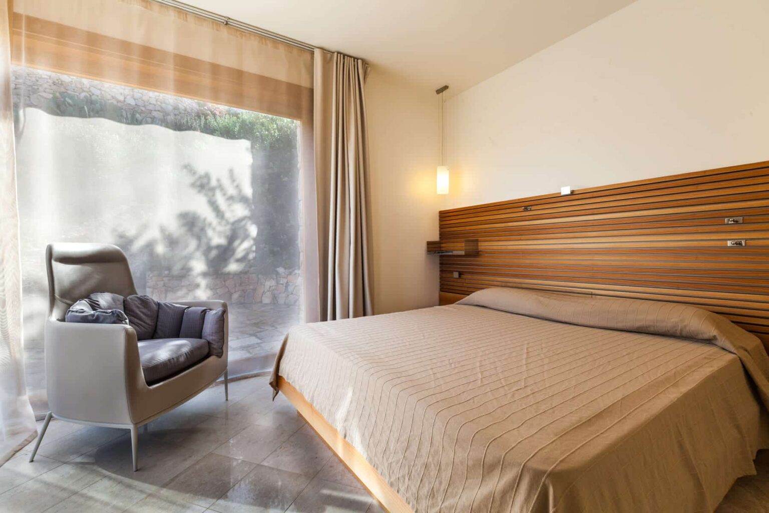 Modern bedroom featuring a queen-sized bed with a beige coverlet, a stylish gray armchair, and large windows with sheer curtains allowing natural light to fill the space. The wooden accent wall adds warmth to the contemporary decor. | Only on SopranoVillas