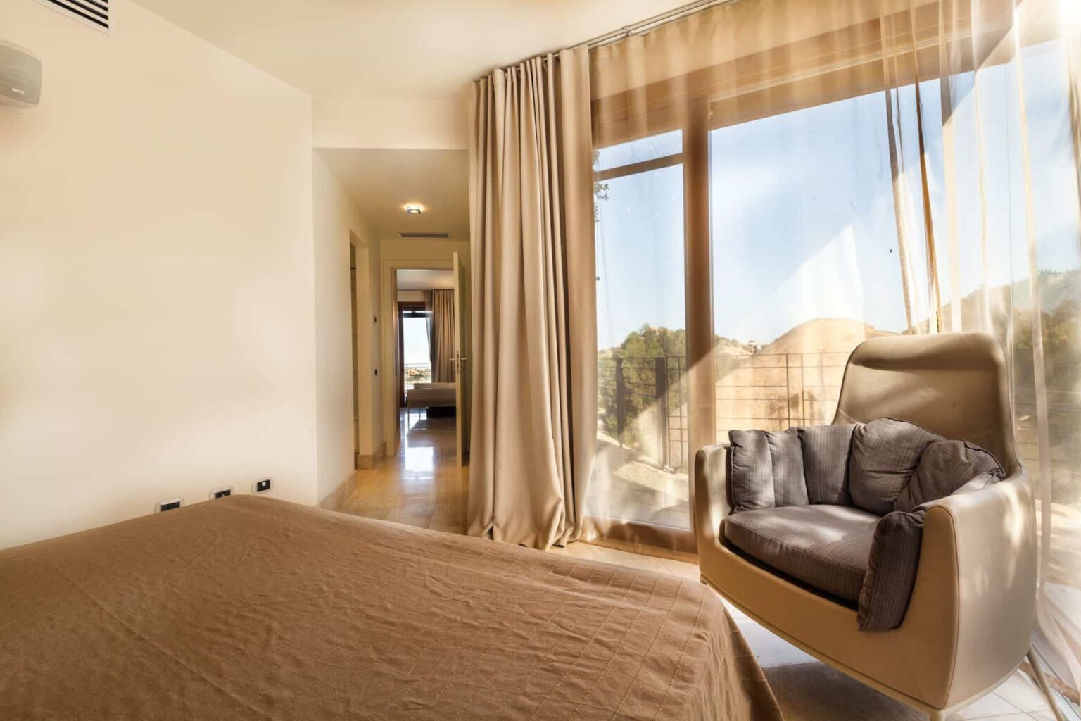 Modern bedroom interior featuring a cozy bed with a brown bedspread, a comfortable armchair by large windows, and soft natural light illuminating the space. | Only on SopranoVillas
