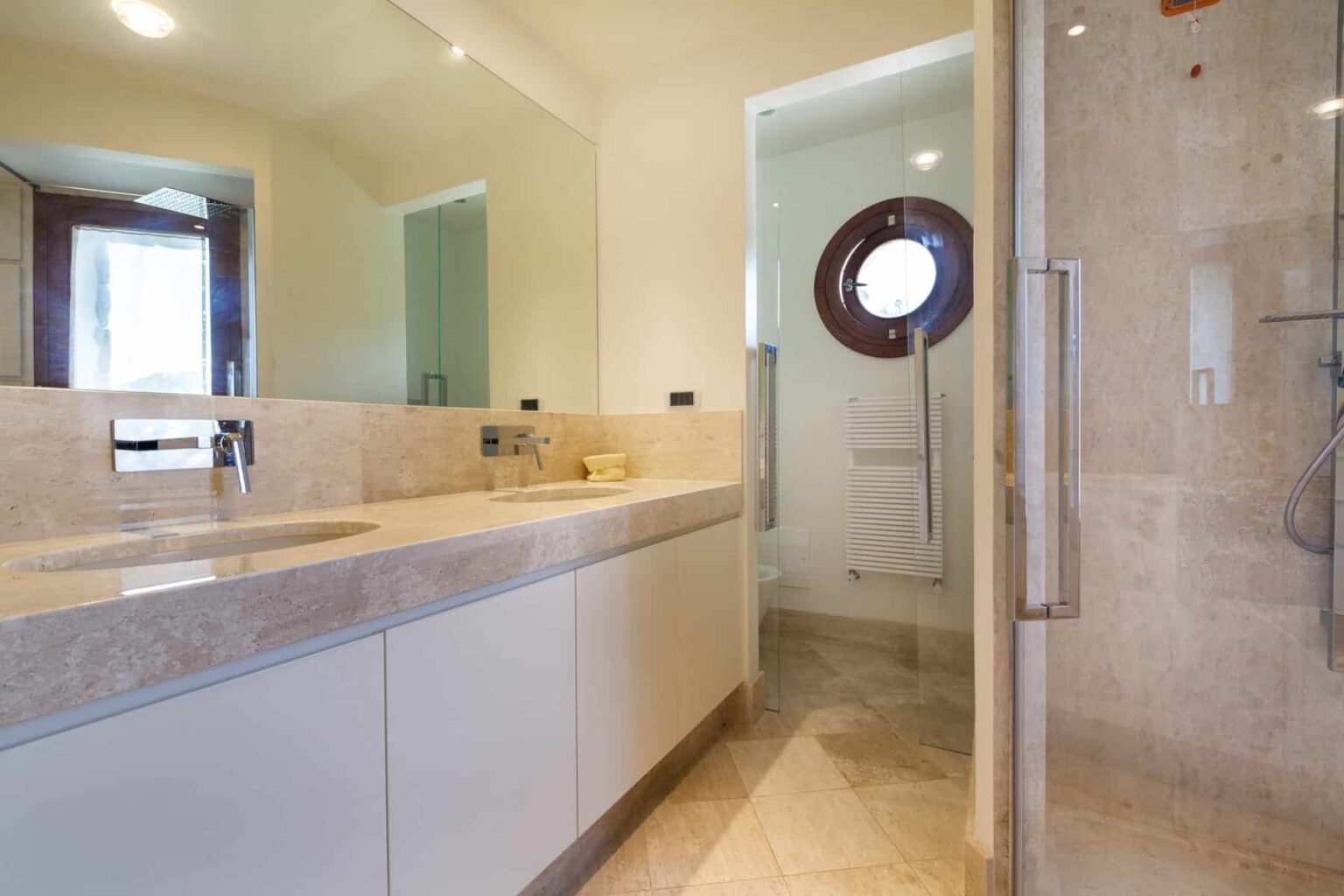 Modern bathroom interior featuring a spacious vanity with a sink, large mirrors, and a glass shower enclosure. Natural light streams in through a circular window, enhancing the elegant design. | Only on SopranoVillas