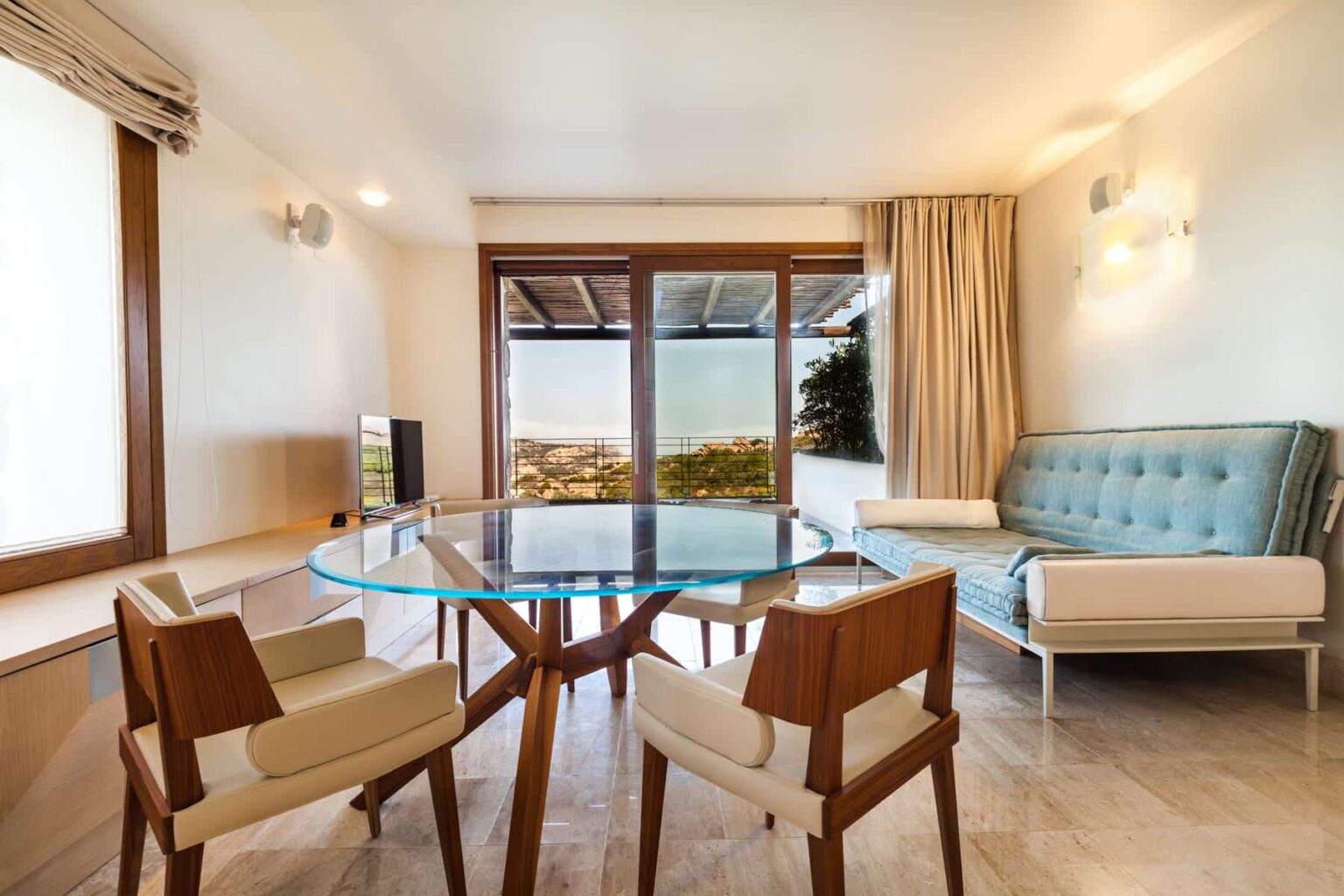 Modern living room with a glass dining table, stylish wooden chairs, and a turquoise sofa, featuring large windows that offer a scenic view. Bright, airy space with neutral decor and natural light. | Only on SopranoVillas