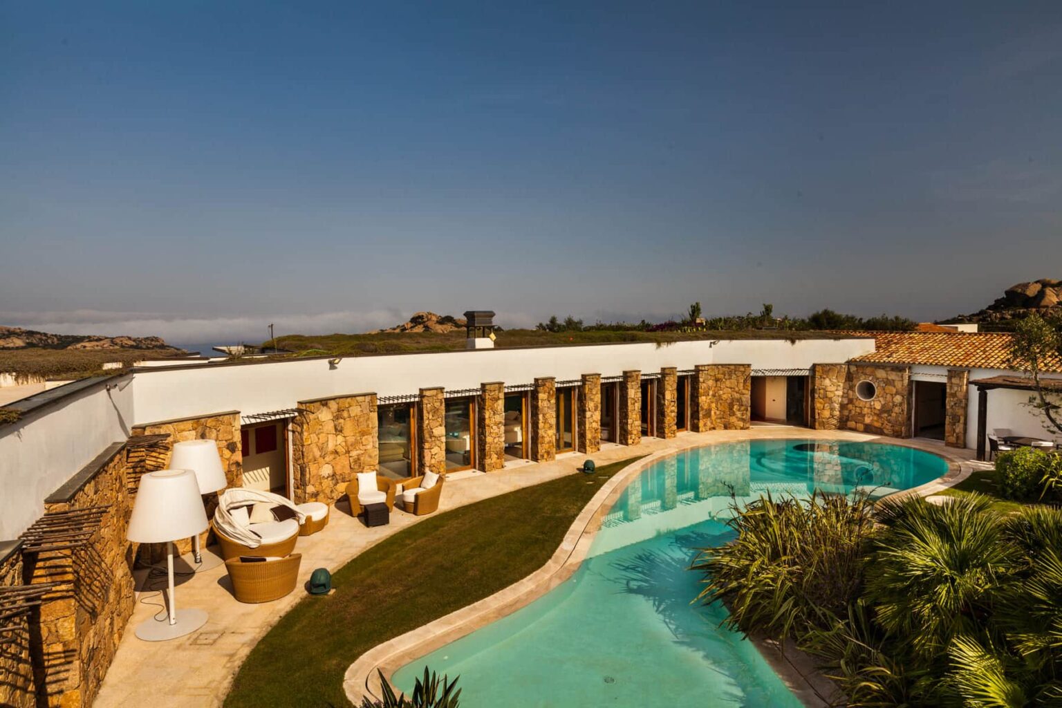Modern luxury villa with a circular design, featuring a serene swimming pool surrounded by lush greenery and stylish outdoor seating areas, set against a clear blue sky. | Only on SopranoVillas