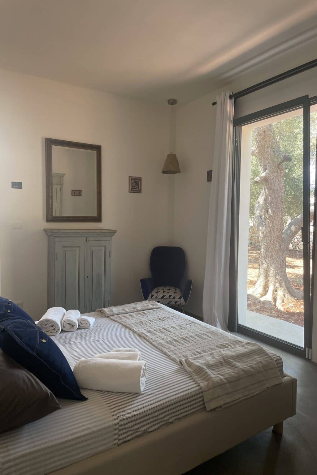 Cozy bedroom interior featuring a neatly made bed with rolled towels, a wooden cabinet, a blue armchair, and a large window that opens to a natural outdoor view. | Only on SopranoVillas