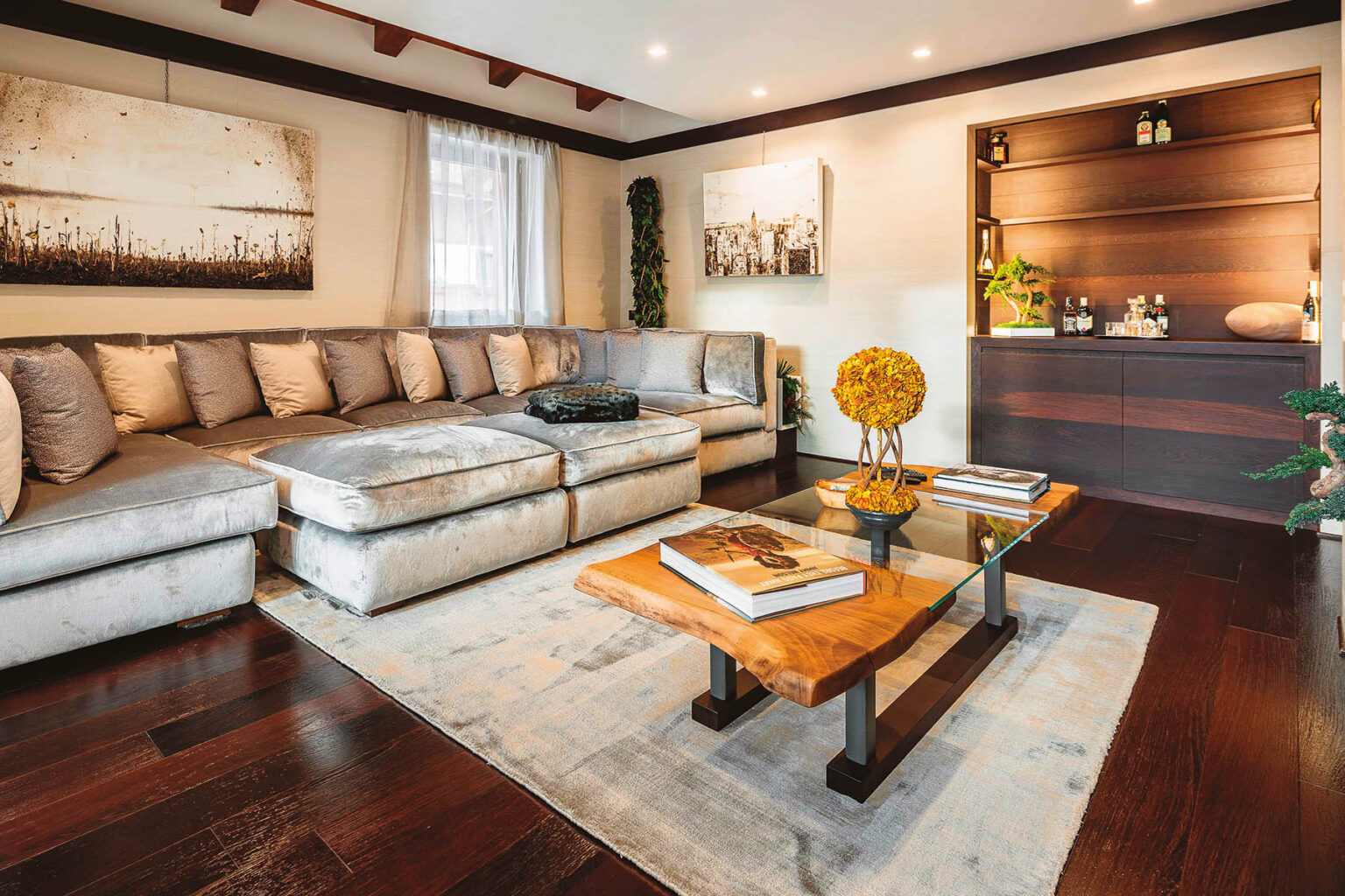 Cozy modern living room featuring a plush sectional sofa with decorative pillows, a wooden coffee table with glass top, and a stylish bar area. The space is illuminated with recessed lighting and natural light from a window, complemented by artwork on the walls and a decorative plant. | Only on SopranoVillas