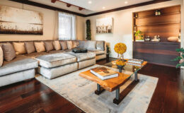 Cozy modern living room featuring a plush sectional sofa with decorative pillows, a wooden coffee table with glass top, and a stylish bar area. The space is illuminated with recessed lighting and natural light from a window, complemented by artwork on the walls and a decorative plant.