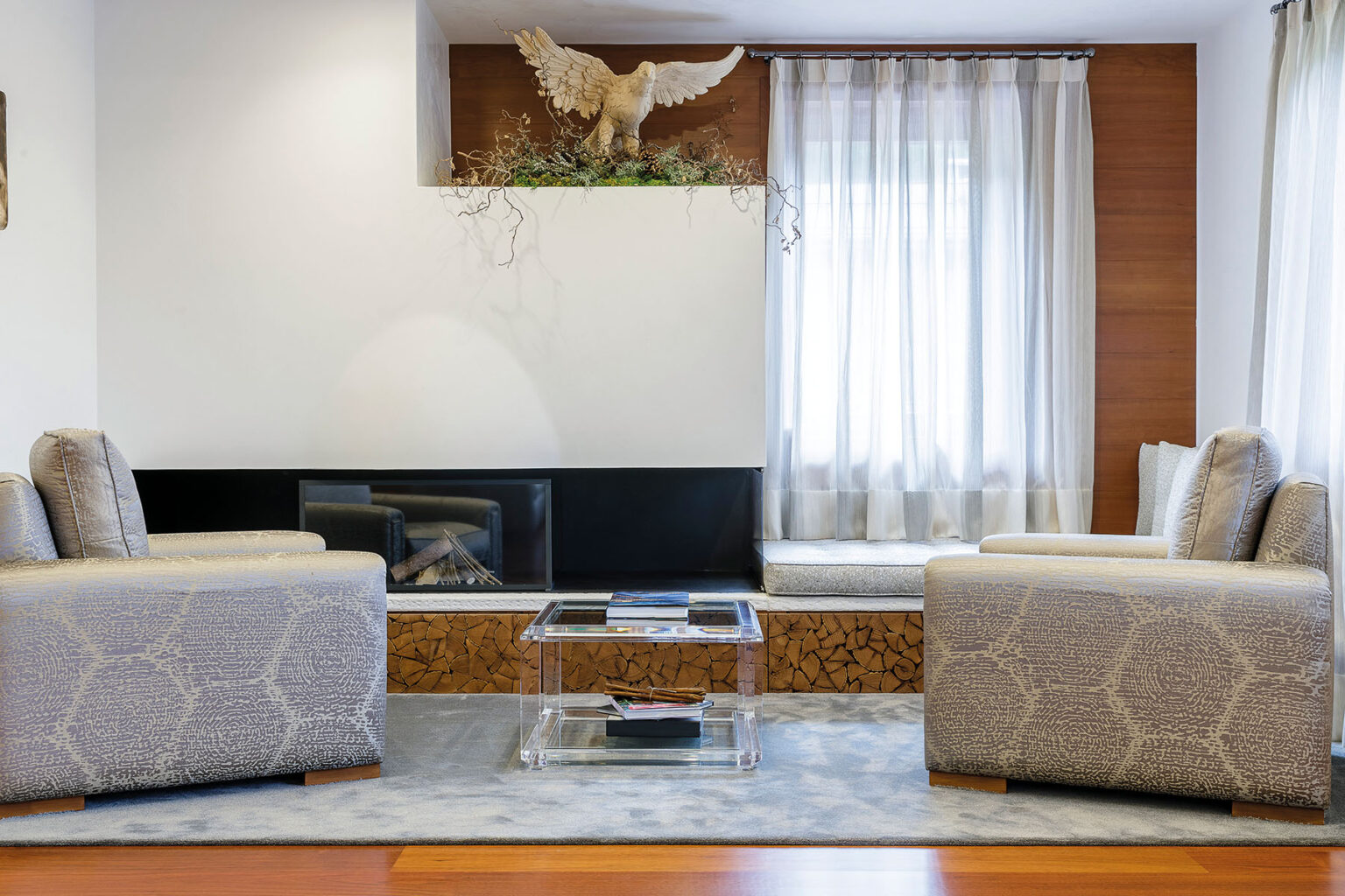 Modern living room featuring two stylish armchairs, a glass coffee table, and large windows allowing natural light. The decor includes a decorative wall with an eagle motif and a cozy fireplace, creating a warm and inviting atmosphere. | Only on SopranoVillas