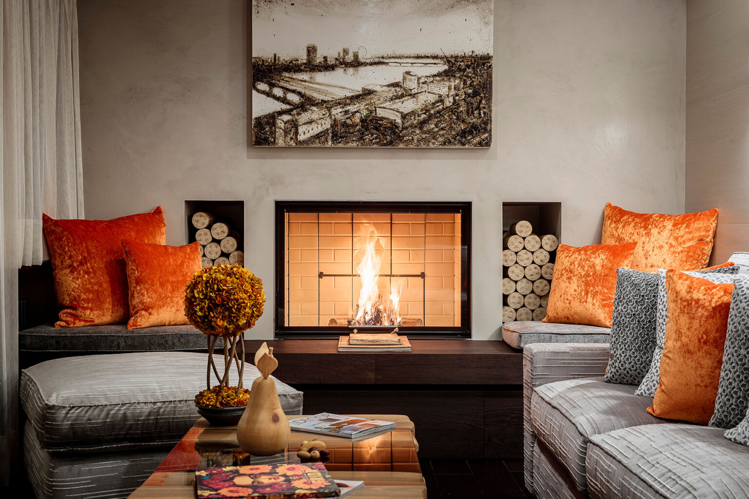Cozy living room with a modern fireplace, featuring orange velvet pillows, a decorative flower arrangement, and a wooden coffee table with magazines. The room has a warm ambiance, highlighted by artwork on the wall and logs neatly stored beside the fireplace. | Only on SopranoVillas