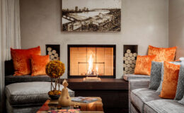 Cozy living room with a modern fireplace, featuring orange velvet pillows, a decorative flower arrangement, and a wooden coffee table with magazines. The room has a warm ambiance, highlighted by artwork on the wall and logs neatly stored beside the fireplace.