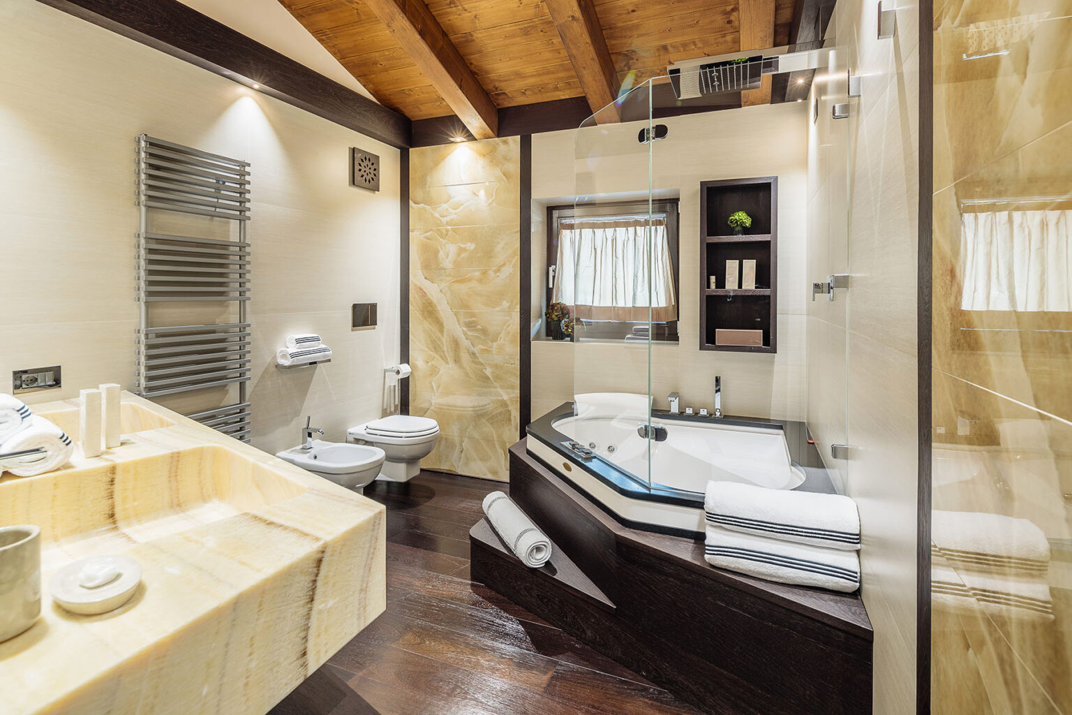 Modern bathroom featuring a luxurious jacuzzi tub, sleek glass shower, stylish wooden beams, and elegant stone finishes. The space includes a spacious sink area with towels and toiletries, creating a serene atmosphere for relaxation. | Only on SopranoVillas
