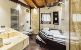 Modern bathroom featuring a luxurious jacuzzi tub, sleek glass shower, stylish wooden beams, and elegant stone finishes. The space includes a spacious sink area with towels and toiletries, creating a serene atmosphere for relaxation.