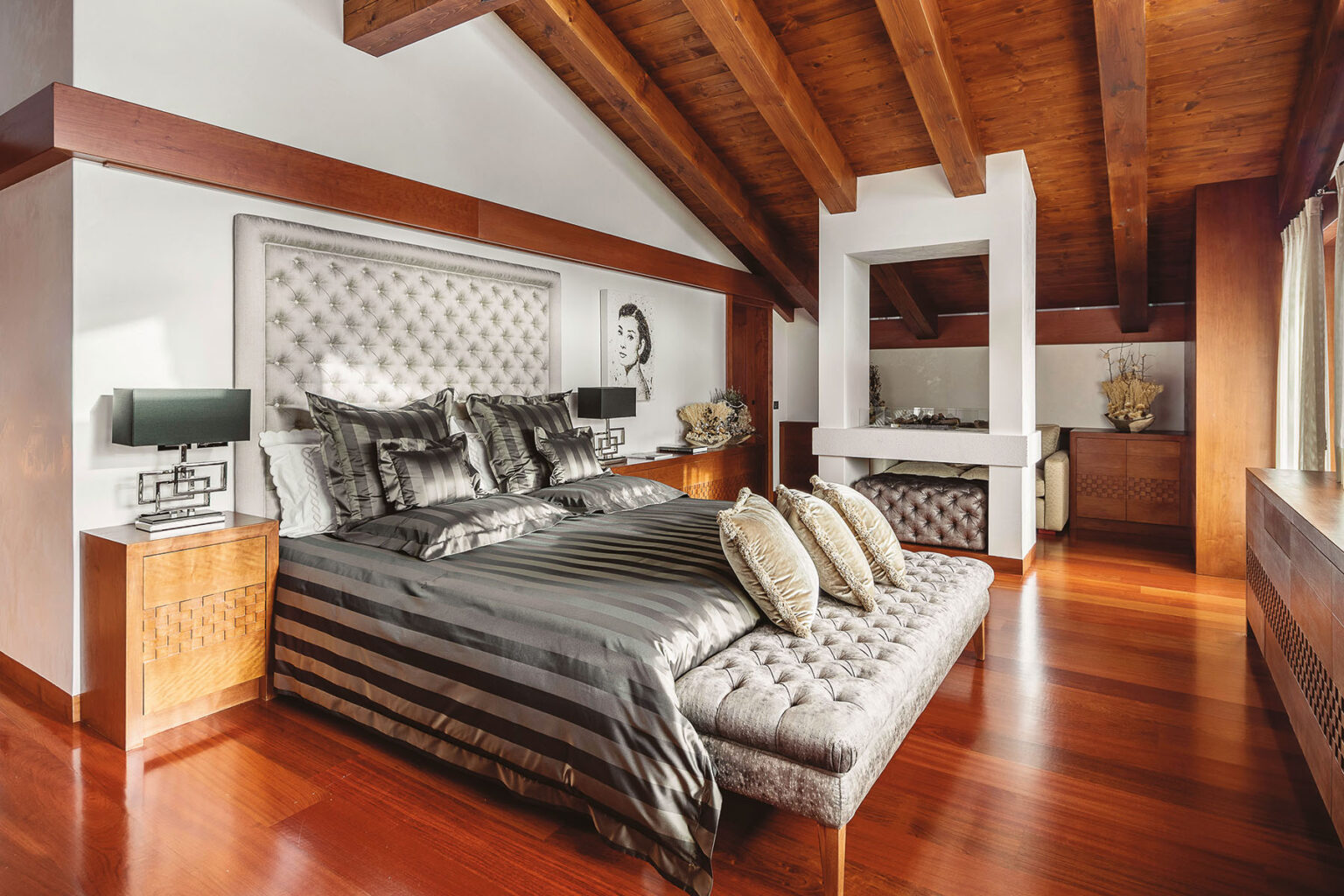 Luxurious modern bedroom with vaulted wooden ceiling, elegant dark bedding, decorative pillows, and stylish wooden furniture, featuring a cozy sitting area and natural light streaming through large windows. | Only on SopranoVillas