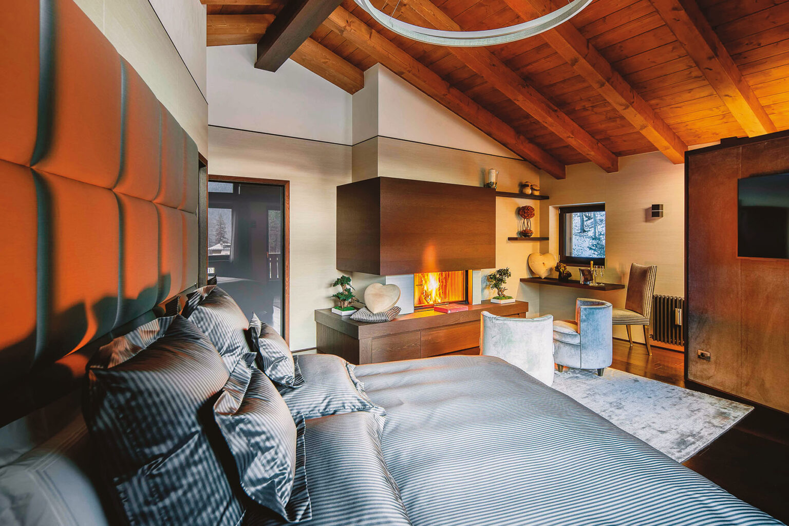 Modern bedroom interior featuring a cozy atmosphere with a king-sized bed adorned with striped pillows, a stylish fireplace integrated into wooden cabinetry, and a workspace with elegant chairs. The room boasts vaulted wooden ceilings and warm lighting, creating a serene retreat. | Only on SopranoVillas