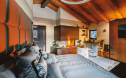 Modern bedroom interior featuring a cozy atmosphere with a king-sized bed adorned with striped pillows, a stylish fireplace integrated into wooden cabinetry, and a workspace with elegant chairs. The room boasts vaulted wooden ceilings and warm lighting, creating a serene retreat.