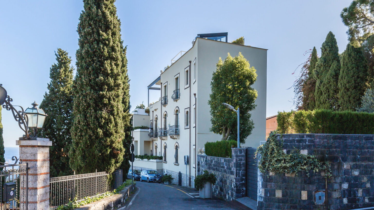 Elegant residential building surrounded by lush greenery and tall cypress trees, featuring a modern architecture design and a serene driveway. Ideal for showcasing luxury real estate or upscale living environments. | Only on SopranoVillas