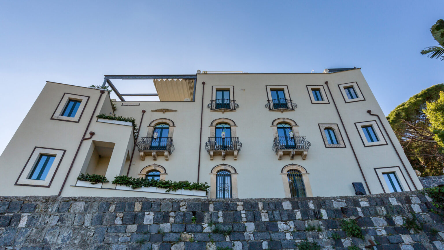 Elegant multi-story building with balconies and a stone wall in the foreground, showcasing modern architecture against a clear blue sky. | Only on SopranoVillas