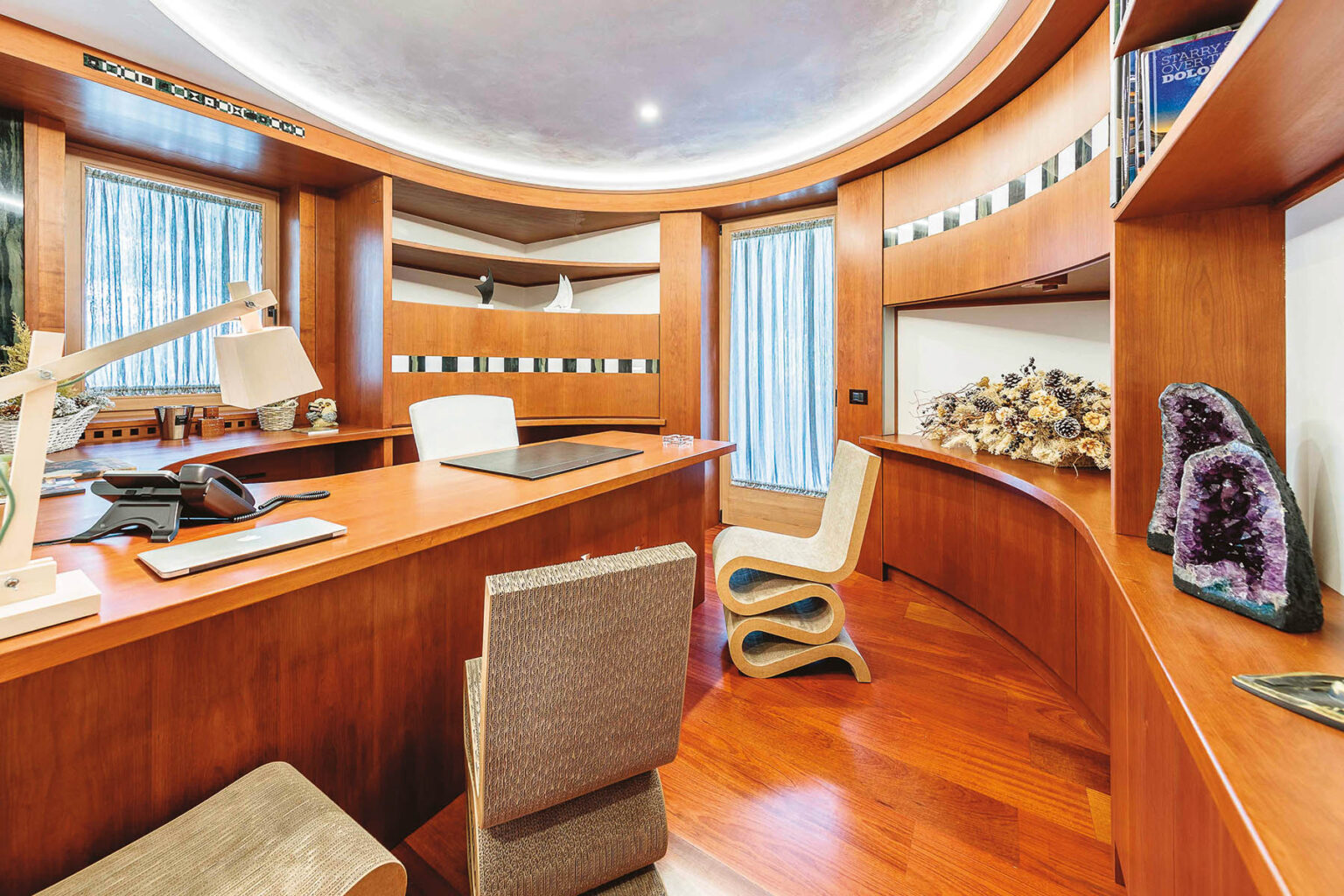 Modern home office interior featuring sleek wooden furniture, a curved desk, and stylish decor elements. The space includes large windows for natural light, a comfortable chair, and decorative items like crystals on the shelves, creating an inviting and functional workspace. | Only on SopranoVillas