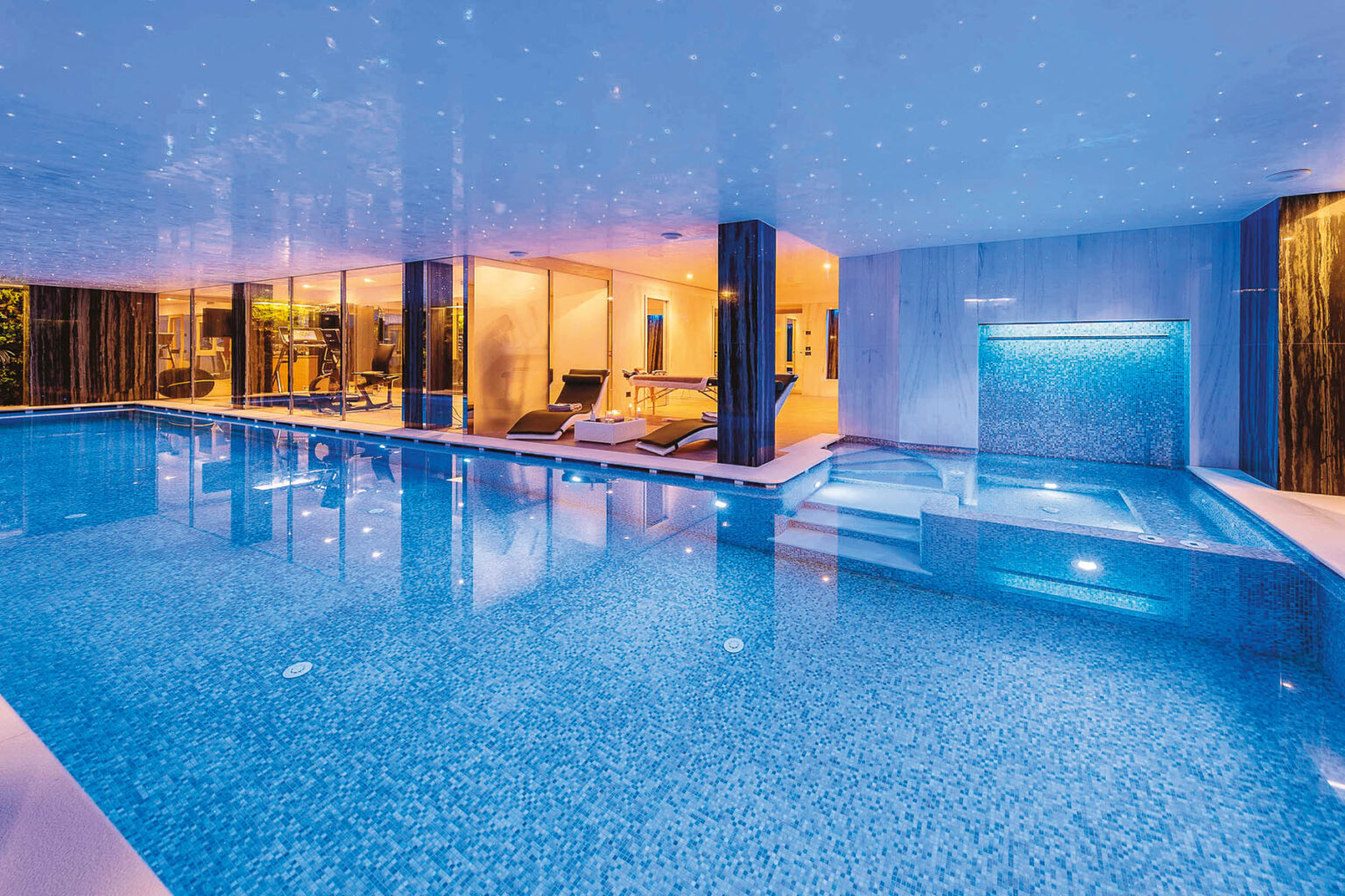Indoor swimming pool with blue mosaic tiles, illuminated by soft lighting and featuring a modern design. The pool area includes lounge chairs and a jacuzzi, surrounded by glass walls that provide a view of the interior space. | Only on SopranoVillas