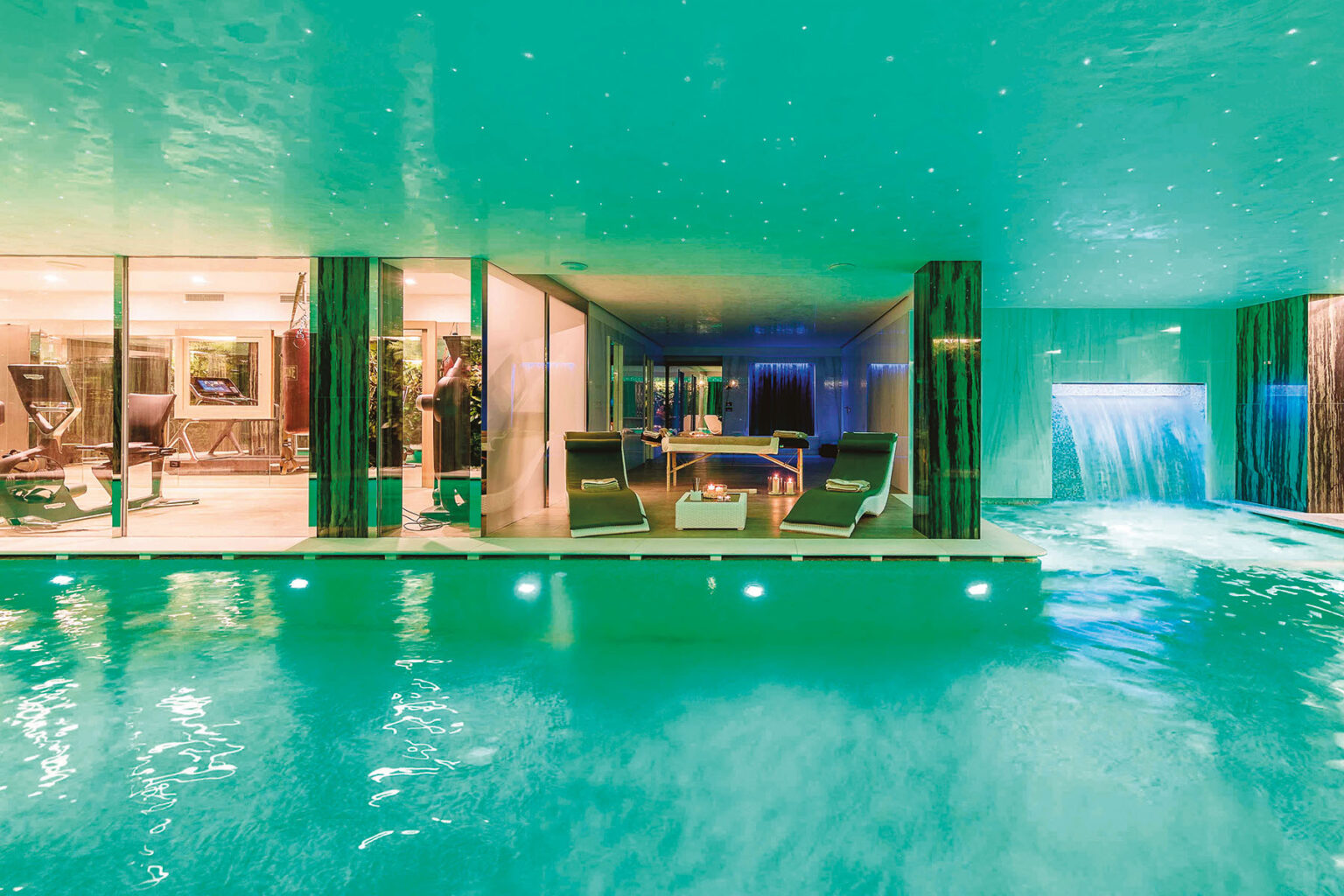 Luxurious indoor pool area featuring modern lounge chairs, a waterfall feature, and a well-equipped fitness space, illuminated with ambient green lighting. Perfect for relaxation and wellness. | Only on SopranoVillas