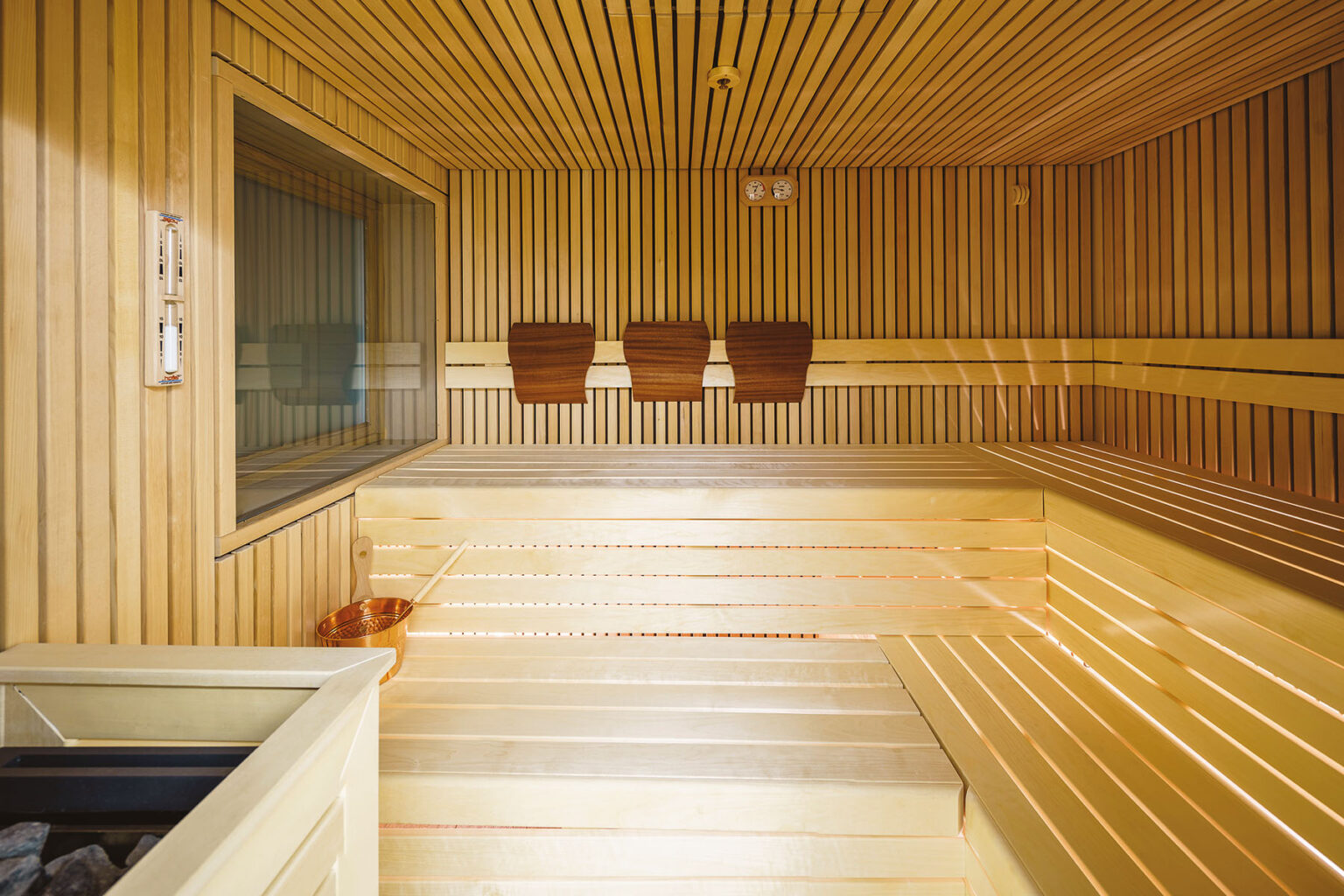Interior of a modern sauna featuring wooden paneling, benches, and a glass window, with a copper bucket and sauna heater visible, creating a relaxing atmosphere for wellness and relaxation. | Only on SopranoVillas