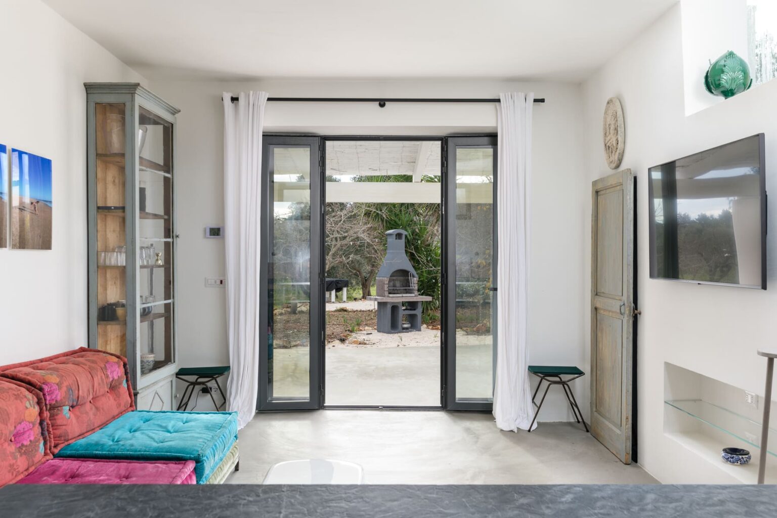 Bright and airy living space featuring large glass doors leading to an outdoor area with a barbecue, colorful sofa, and modern decor. | Only on SopranoVillas