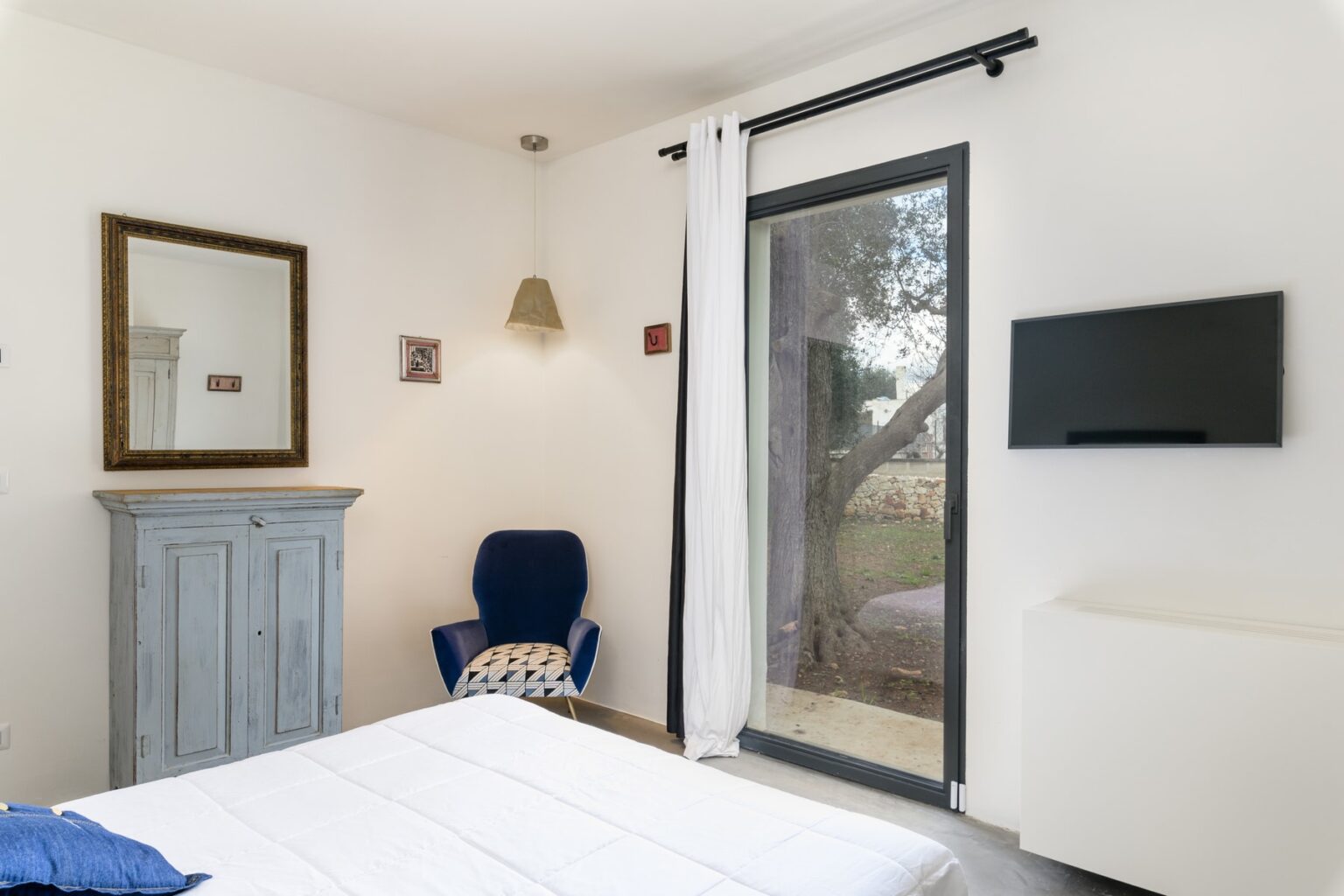 Cozy modern bedroom featuring a white bedspread, a blue chair with geometric patterns, a wooden cabinet, a large mirror, and a mounted TV. Natural light streams in through the glass door, which opens to a scenic outdoor view. Ideal for relaxation and comfort. | Only on SopranoVillas