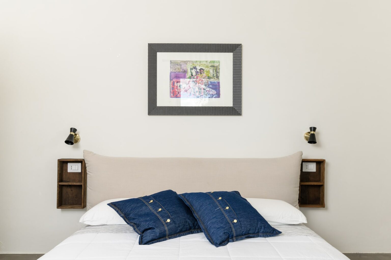 Cozy bedroom featuring a neutral-colored headboard, blue decorative pillows on a white bed, and a framed artwork above. Two bedside lamps and wooden shelves enhance the modern decor. Ideal for home staging or interior design inspiration. | Only on SopranoVillas