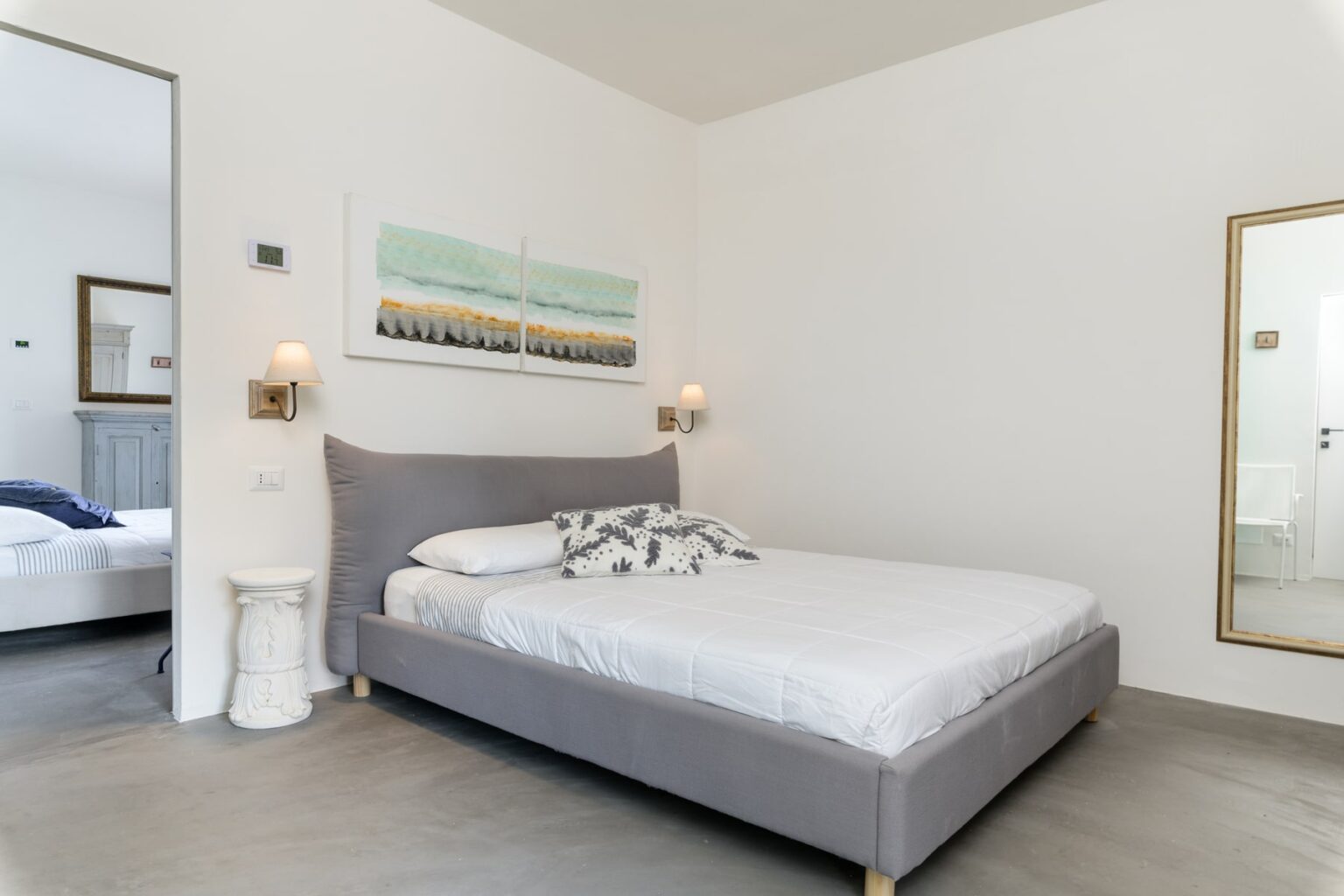 Modern bedroom featuring a gray bed with decorative pillows, minimalist decor, and natural light. The room includes stylish wall art and a large mirror, creating a spacious ambiance. Ideal for relaxation and comfort. | Only on SopranoVillas
