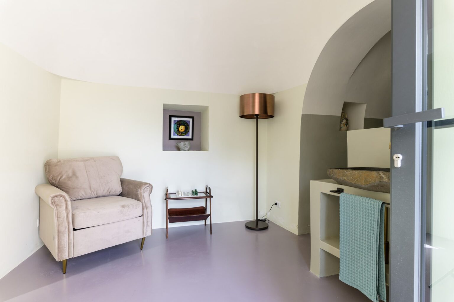 Cozy interior space featuring a light-colored armchair, a modern floor lamp, a small side table, and a minimalist console with a decorative item, set against a softly curved wall in a well-lit room. | Only on SopranoVillas