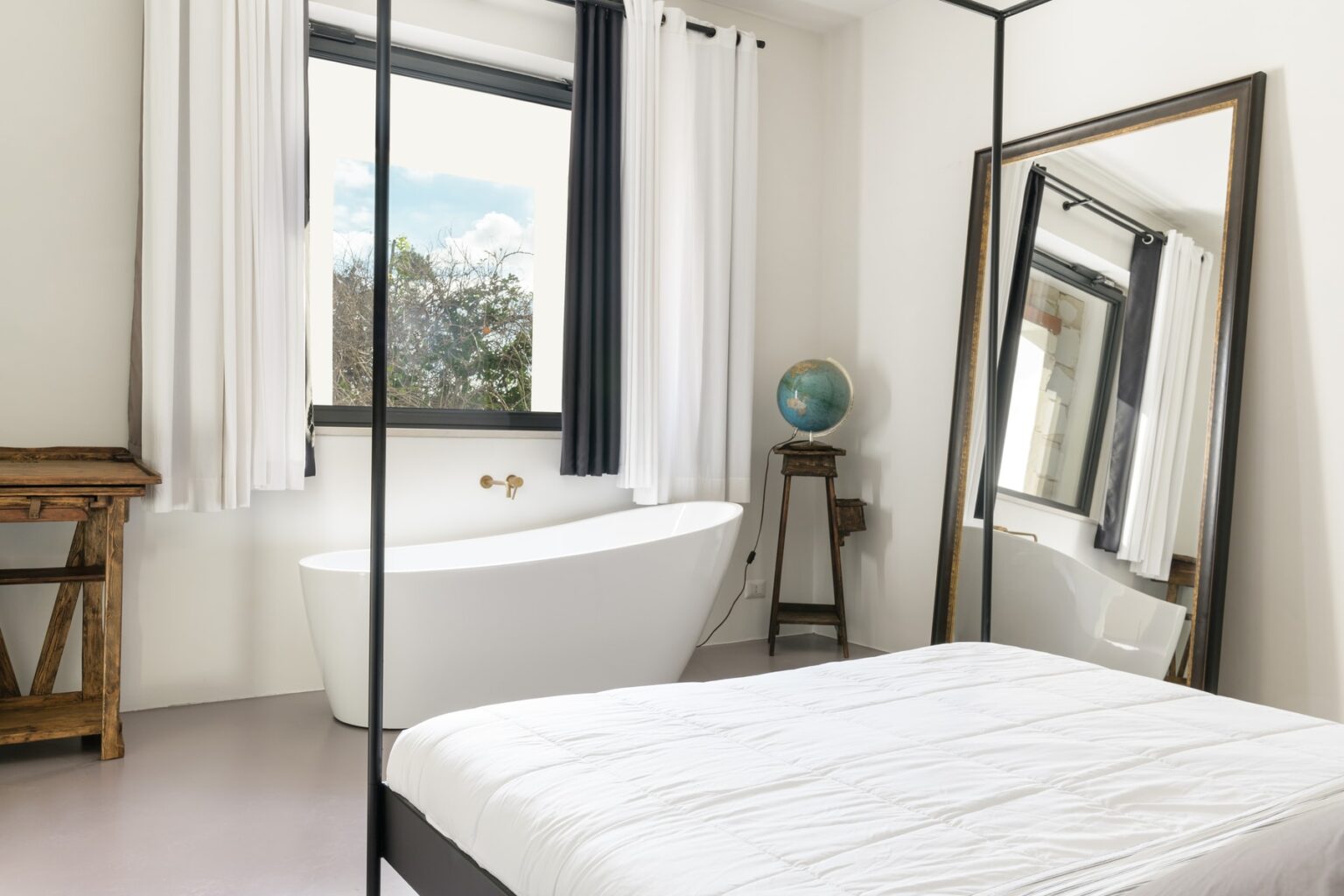 Modern bedroom featuring a white bed, large mirror, and a freestanding bathtub by the window, accented with natural light and minimalist decor. | Only on SopranoVillas