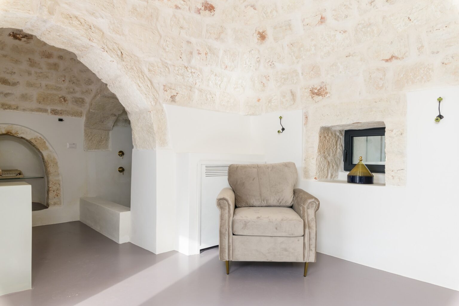 Cozy interior design featuring a plush beige armchair in a modern space with stone archways and a minimalist aesthetic. Natural light highlights the unique architectural details, creating a warm and inviting atmosphere. | Only on SopranoVillas