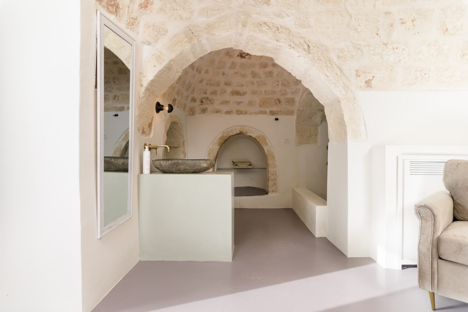 Interior view of a modern, minimalist living space featuring arched stone walls, a contemporary kitchen area with a glass sink, and a cozy seating area. The design blends historical architecture with modern furnishings, creating a stylish and inviting atmosphere. | Only on SopranoVillas