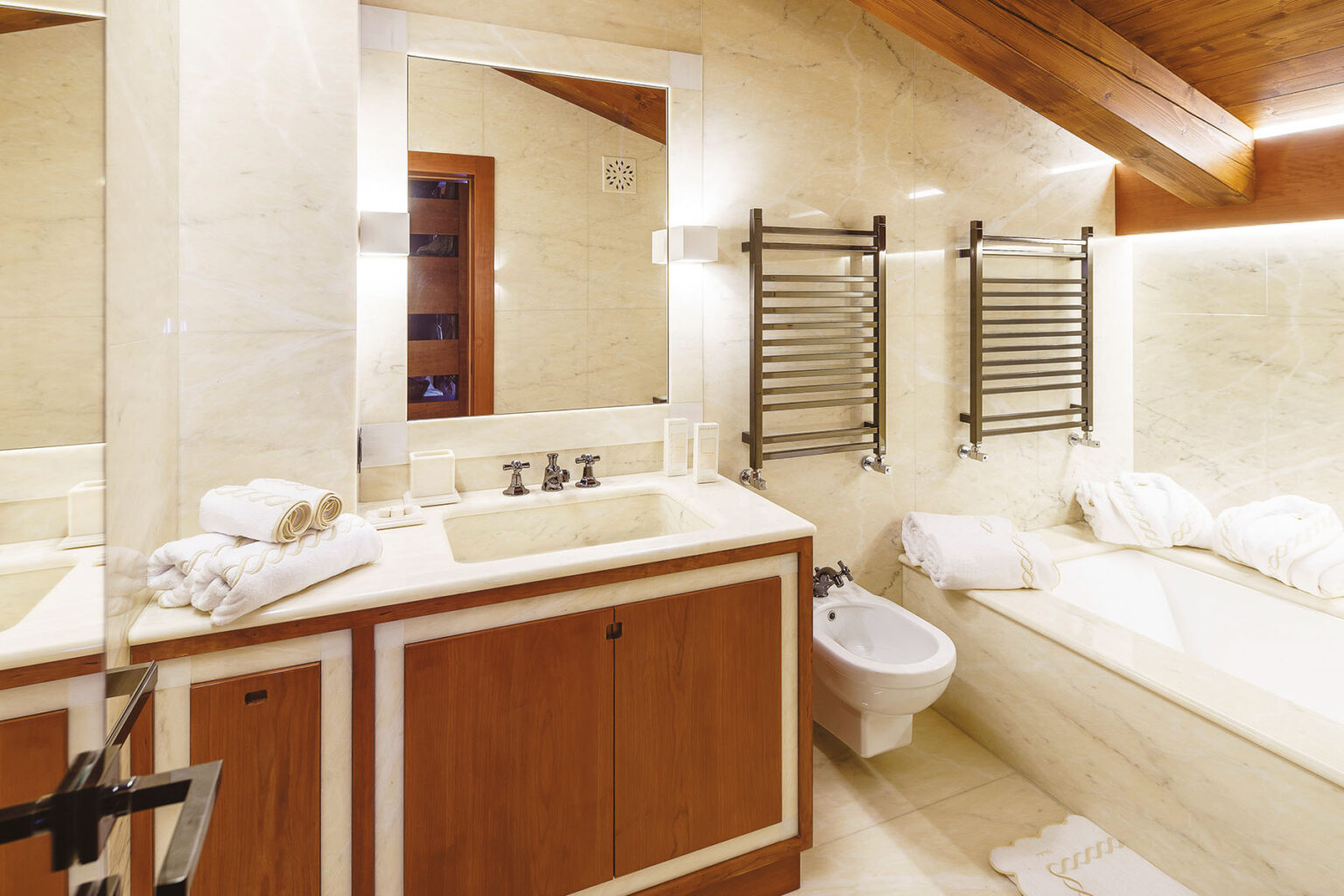 Modern bathroom interior featuring a marble finish, wooden cabinetry, a double sink vanity with stylish faucets, a bidet, and a bathtub. The space is well-lit with wall-mounted lights and includes neatly arranged white towels for a luxurious touch. | Only on SopranoVillas