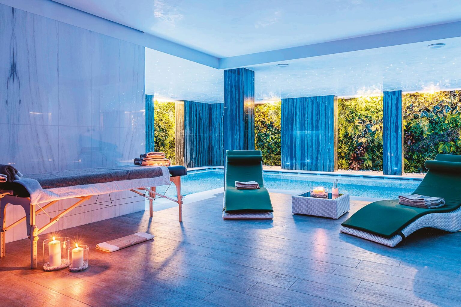 Serene spa interior featuring a massage table, two green lounge chairs, and a tranquil pool surrounded by lush greenery and soft candlelight for a relaxing atmosphere. | Only on SopranoVillas