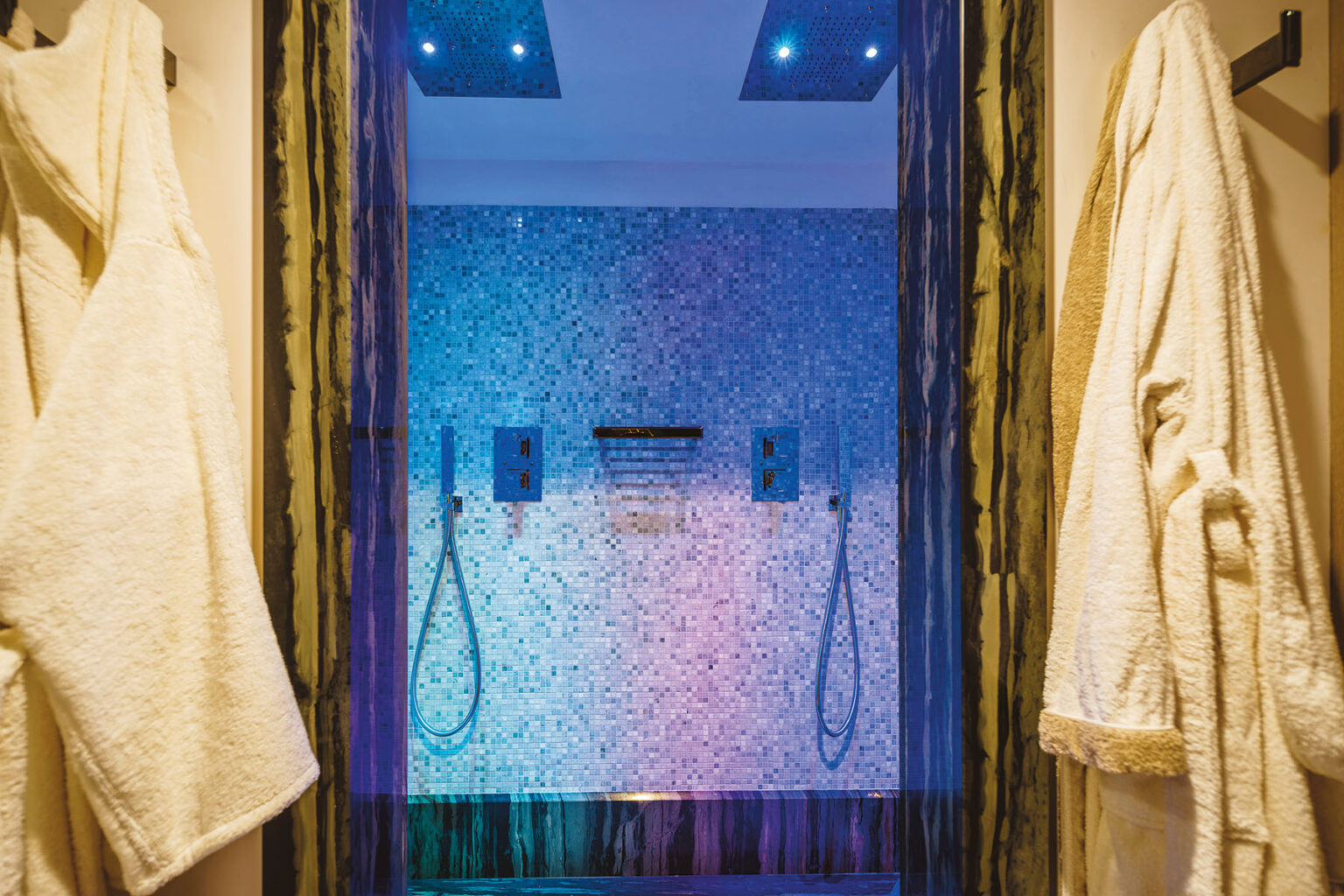 Luxurious modern shower featuring a mosaic tile wall in shades of blue and purple, equipped with dual showerheads and stylish fixtures, with plush white bathrobes hanging on the side. | Only on SopranoVillas