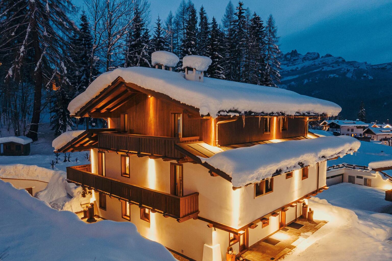 Cozy chalet covered in snow, illuminated by warm lights, surrounded by tall evergreen trees during twilight in a winter landscape. | Only on SopranoVillas