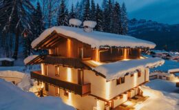 Cozy chalet covered in snow, illuminated by warm lights, surrounded by tall evergreen trees during twilight in a winter landscape.