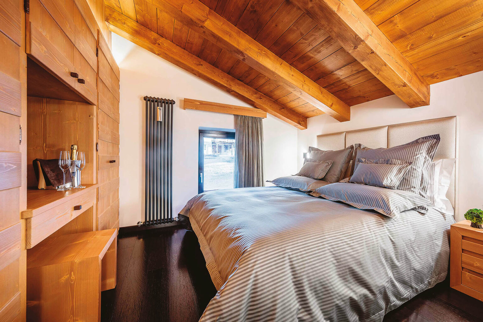 Cozy bedroom featuring a wooden ceiling with exposed beams, a comfortable bed with gray bedding, and stylish wooden furniture, including a nightstand and storage cabinet, creating a warm and inviting atmosphere. | Only on SopranoVillas