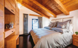 Cozy bedroom featuring a wooden ceiling with exposed beams, a comfortable bed with gray bedding, and stylish wooden furniture, including a nightstand and storage cabinet, creating a warm and inviting atmosphere.