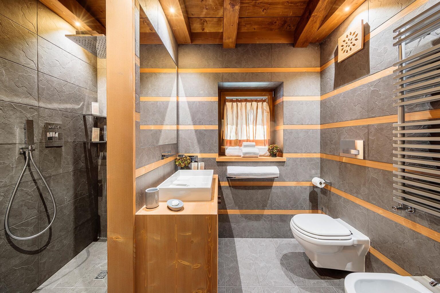 Modern bathroom interior featuring a combination of wooden beams and gray stone walls, equipped with a walk-in shower, sleek vanity, and a toilet, illuminated by natural light from a window. Ideal for showcasing contemporary design and functionality. | Only on SopranoVillas