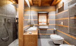 Modern bathroom interior featuring a combination of wooden beams and gray stone walls, equipped with a walk-in shower, sleek vanity, and a toilet, illuminated by natural light from a window. Ideal for showcasing contemporary design and functionality.