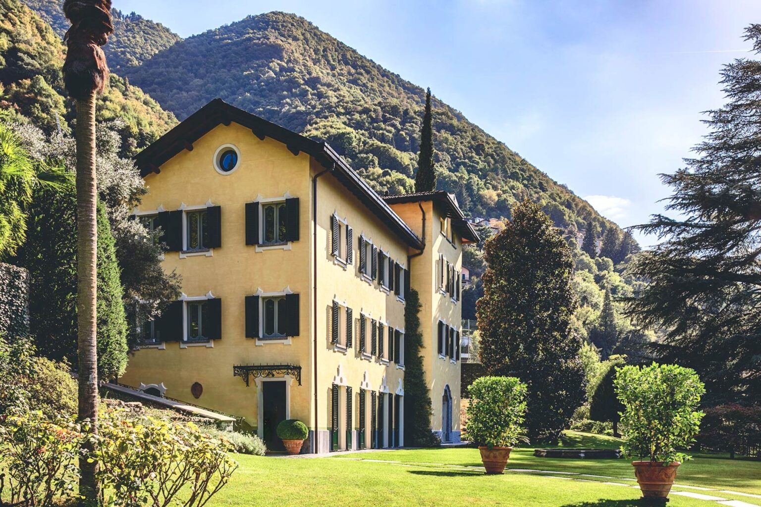 Historic villa surrounded by lush greenery and mountains, featuring a yellow facade and large windows, set in a picturesque landscape. | Only on SopranoVillas
