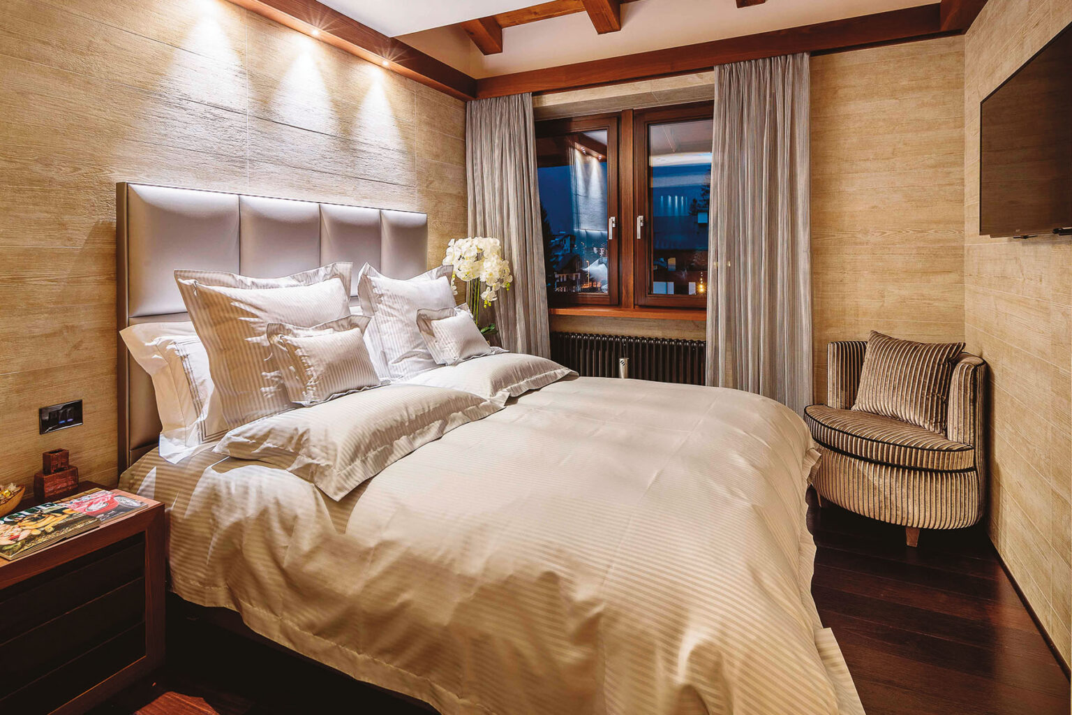 Cozy and elegant bedroom featuring a plush bed with soft bedding, stylish headboard, and ambient lighting. The room includes large windows with scenic views, tasteful curtains, and a comfortable armchair, creating a welcoming atmosphere for relaxation. | Only on SopranoVillas