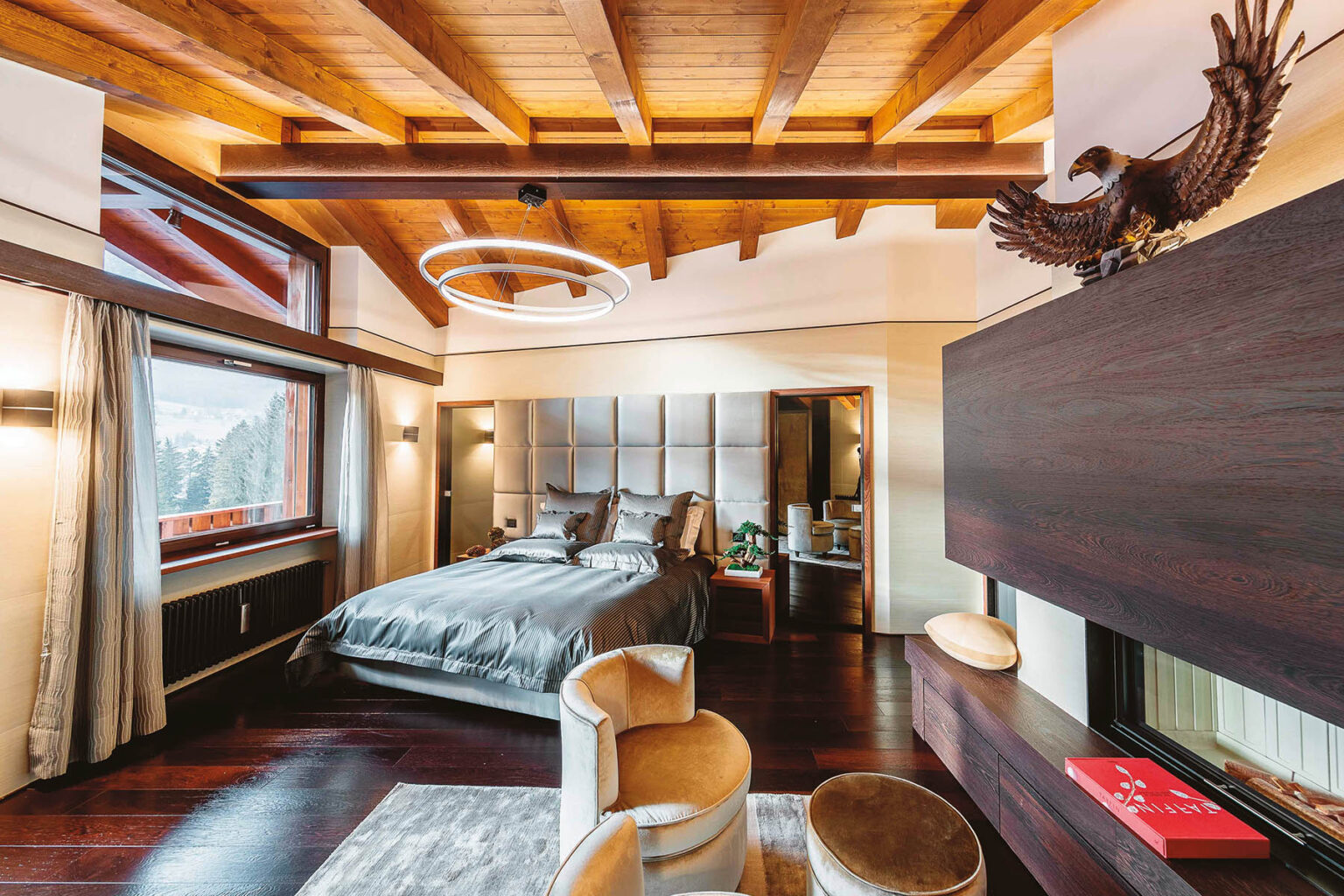 Modern bedroom design featuring a luxurious bed with gray bedding, wooden ceiling beams, and large windows showcasing a scenic view. The room includes stylish accent chairs, contemporary lighting, and elegant decor elements, creating a cozy and inviting atmosphere. | Only on SopranoVillas