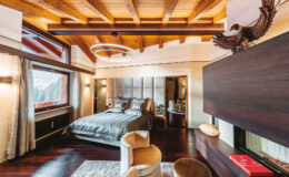 Modern bedroom design featuring a luxurious bed with gray bedding, wooden ceiling beams, and large windows showcasing a scenic view. The room includes stylish accent chairs, contemporary lighting, and elegant decor elements, creating a cozy and inviting atmosphere.