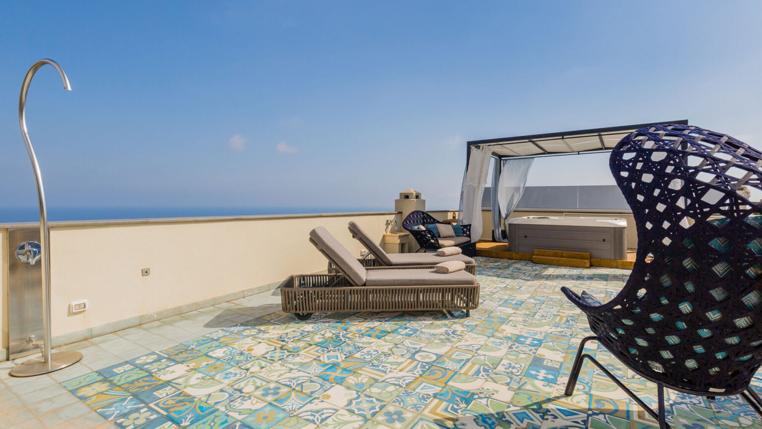 Rooftop terrace with lounge chairs, a shaded seating area, and a hot tub, overlooking a clear blue sky and ocean view. The floor features colorful tiles, creating a vibrant outdoor space for relaxation. | Only on SopranoVillas