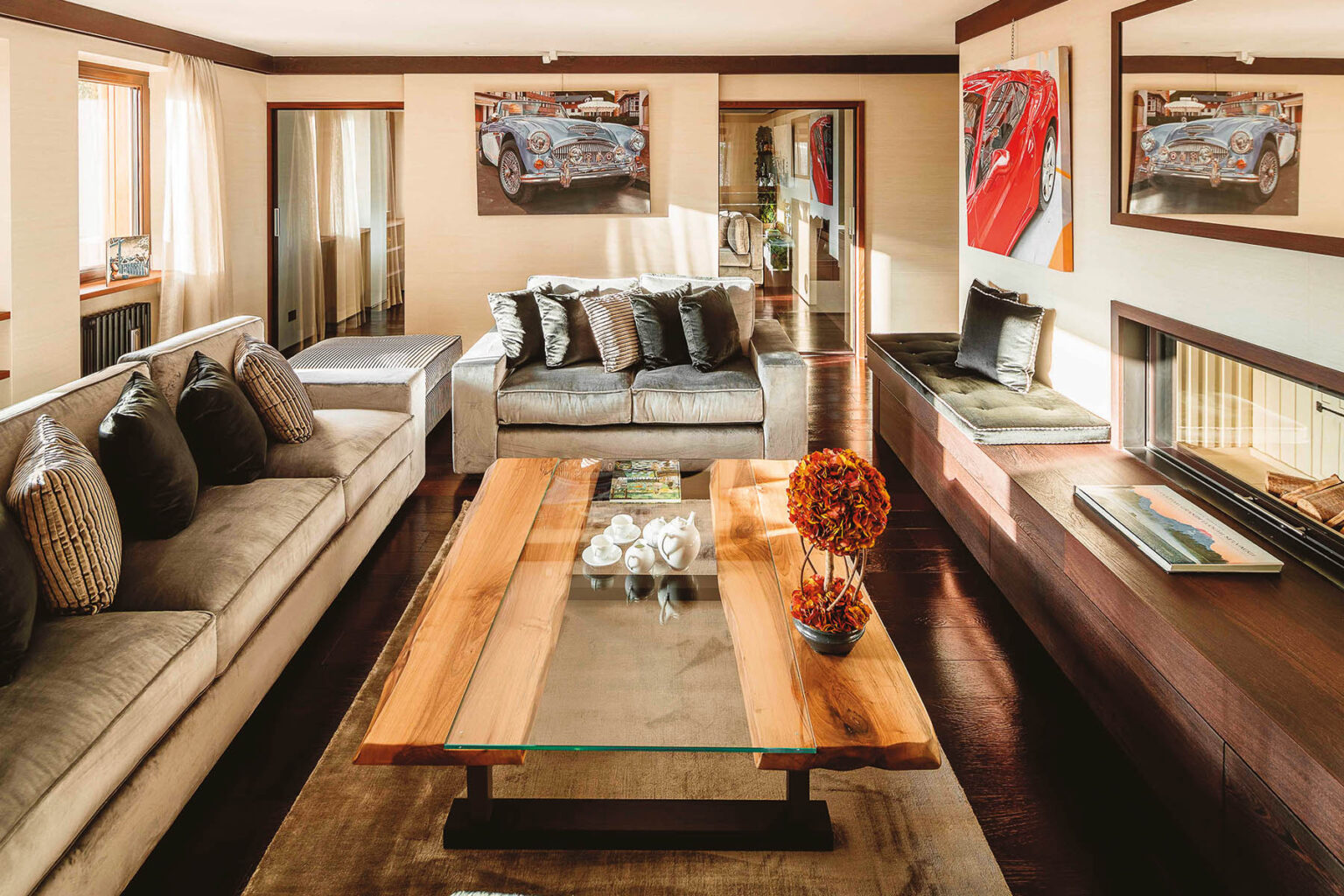 Modern living room featuring a stylish design with a gray sectional sofa, decorative pillows, and a wooden coffee table with a glass top. The room is adorned with artwork of classic cars and has large windows that allow natural light to illuminate the space. A cozy seating area is created near a contemporary fireplace, enhancing the inviting atmosphere. | Only on SopranoVillas