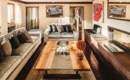 Modern living room featuring a stylish design with a gray sectional sofa, decorative pillows, and a wooden coffee table with a glass top. The room is adorned with artwork of classic cars and has large windows that allow natural light to illuminate the space. A cozy seating area is created near a contemporary fireplace, enhancing the inviting atmosphere.