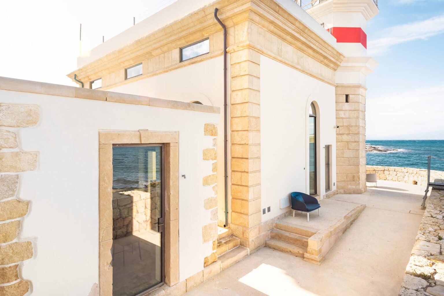Exterior view of a coastal lighthouse featuring a stone facade, large windows, and a red and white striped top, with a scenic ocean backdrop. | Only on SopranoVillas